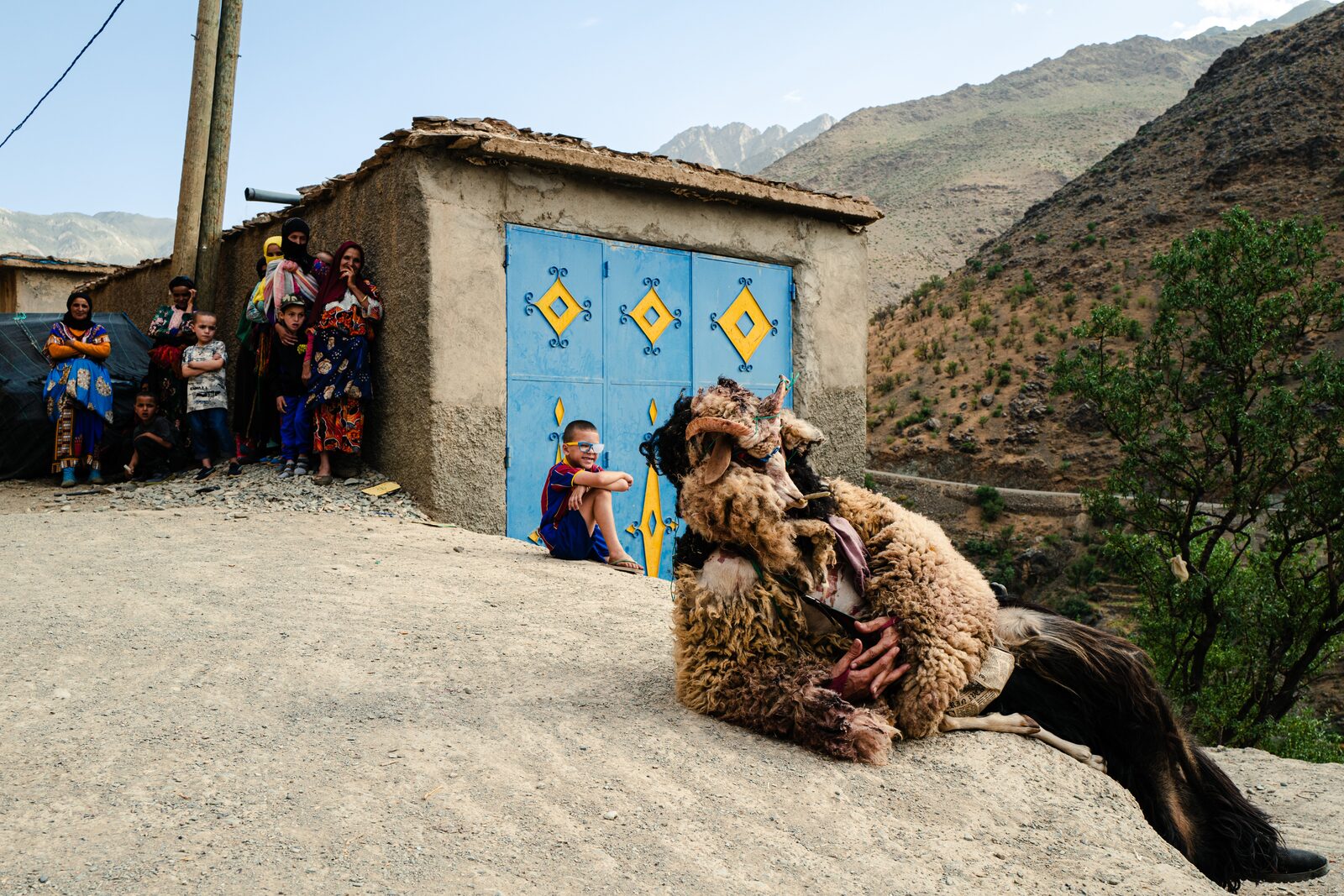 Boujloud performer in sheepskin lying on a dusty slope before a house with blue doors, mountains behind — Abderrahmane Ajja for ZELZAL