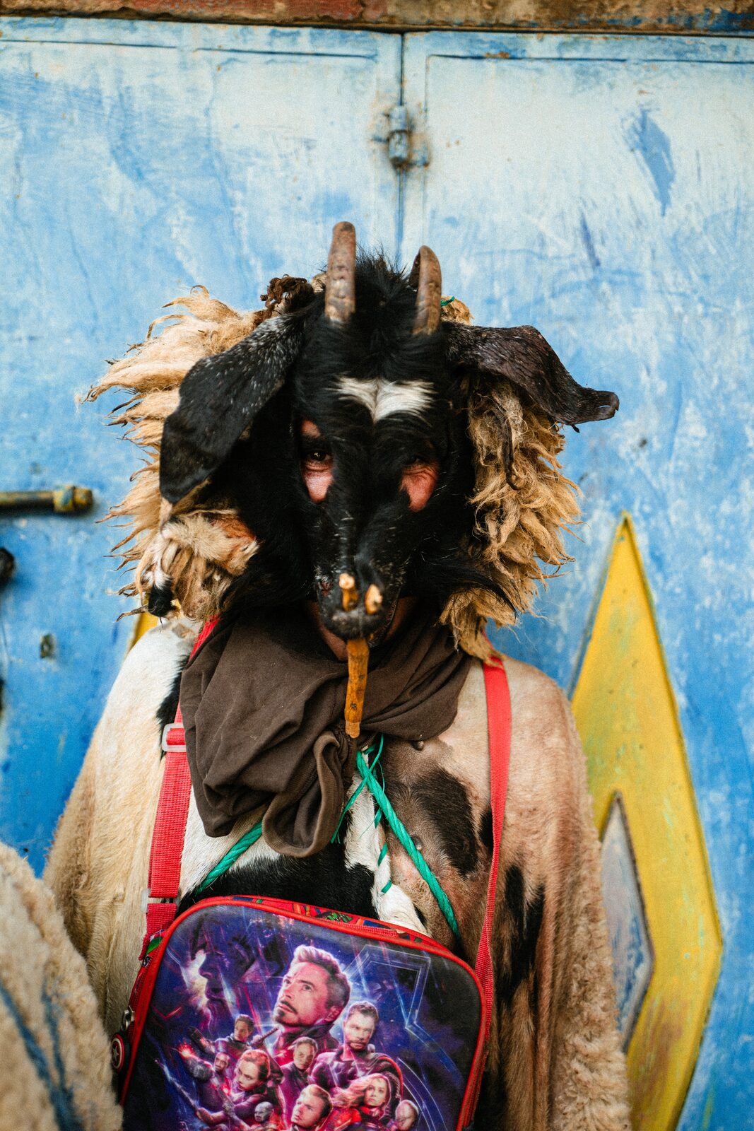 Boujloud figure in a horned animal-skin mask against a blue wall — Abderrahmane Ajja for ZELZAL