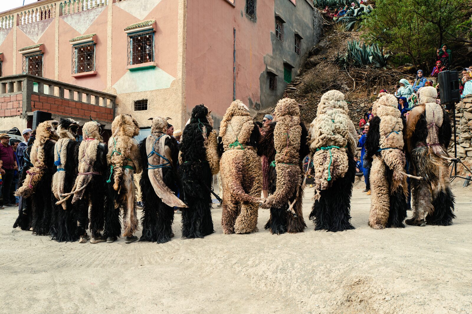 Crowd of Boujloud performers in sheepskin costumes outside a pink village house — Abderrahmane Ajja for ZELZAL