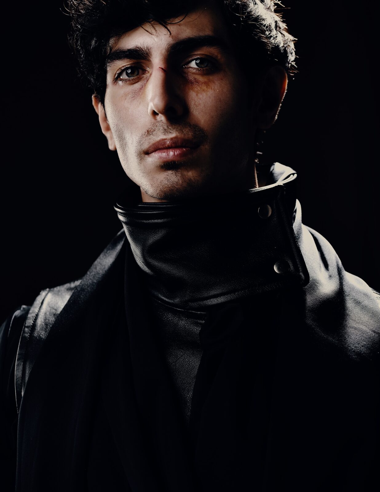 Young man in a black high-collared coat, lit against a black background — Abdullah Al Zanki for ZELZAL
