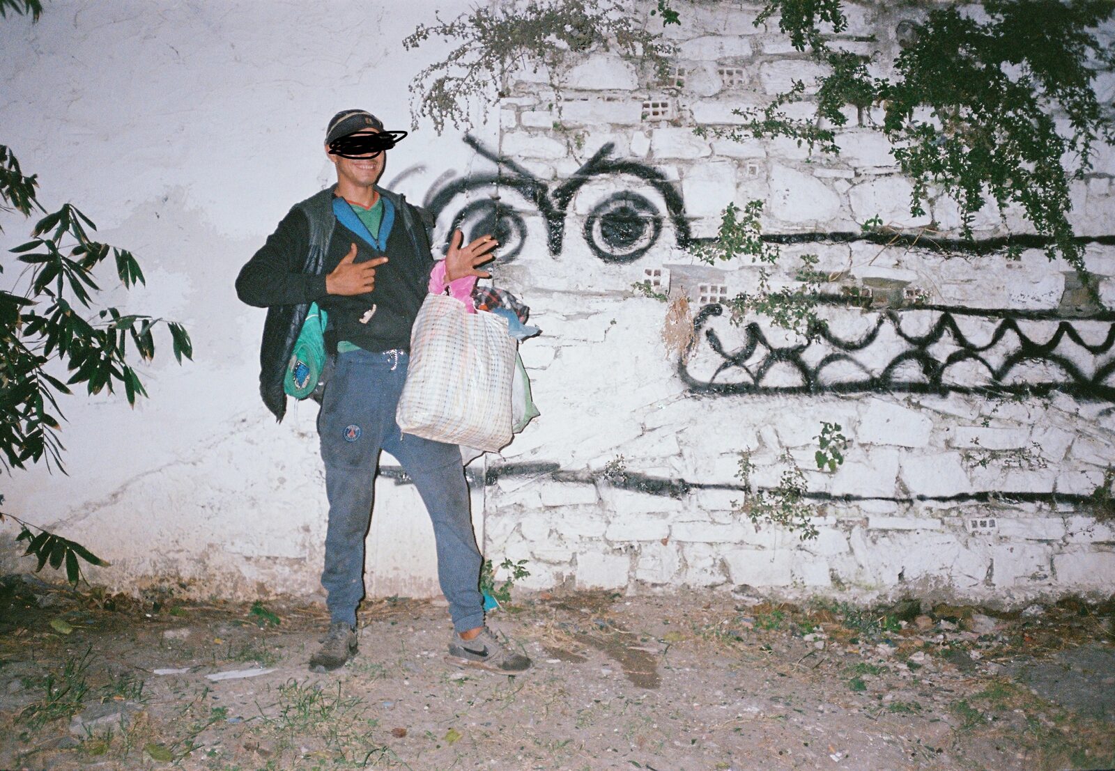 Man in a cap holding a bag in front of a wall painted with monster eyes — Bluuecrab for ZELZAL