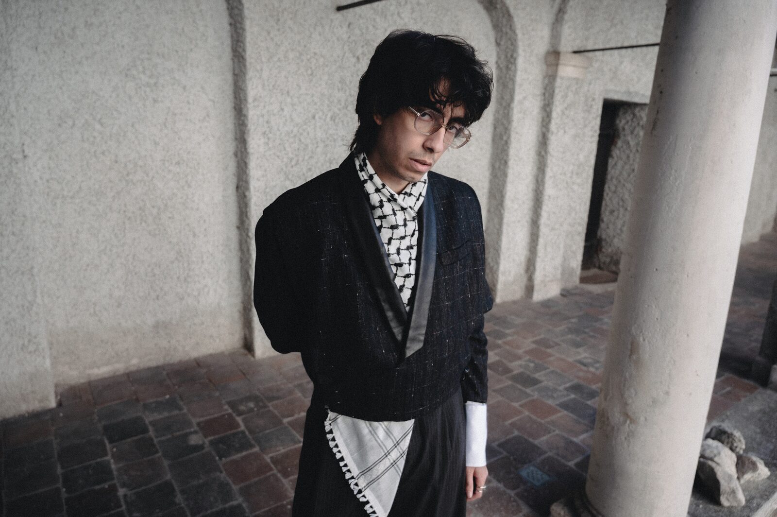Young man in glasses, black jacket and keffiyeh scarf beside a stone column — Deza for ZELZAL