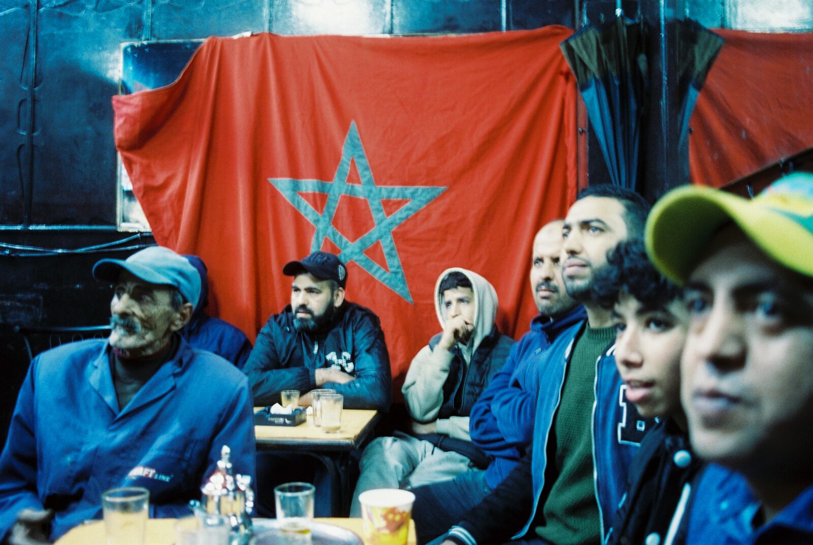 Men at a cafe table in front of a Moroccan flag — Ilias El Faris for ZELZAL