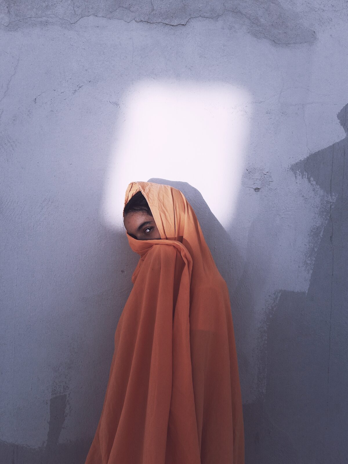 Woman veiled in terracotta fabric beneath a square of light on a wall — Ismail Zaidy for ZELZAL