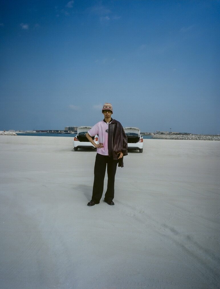 Man in a pink shirt standing on a pale empty lot by a car — Moz for ZELZAL