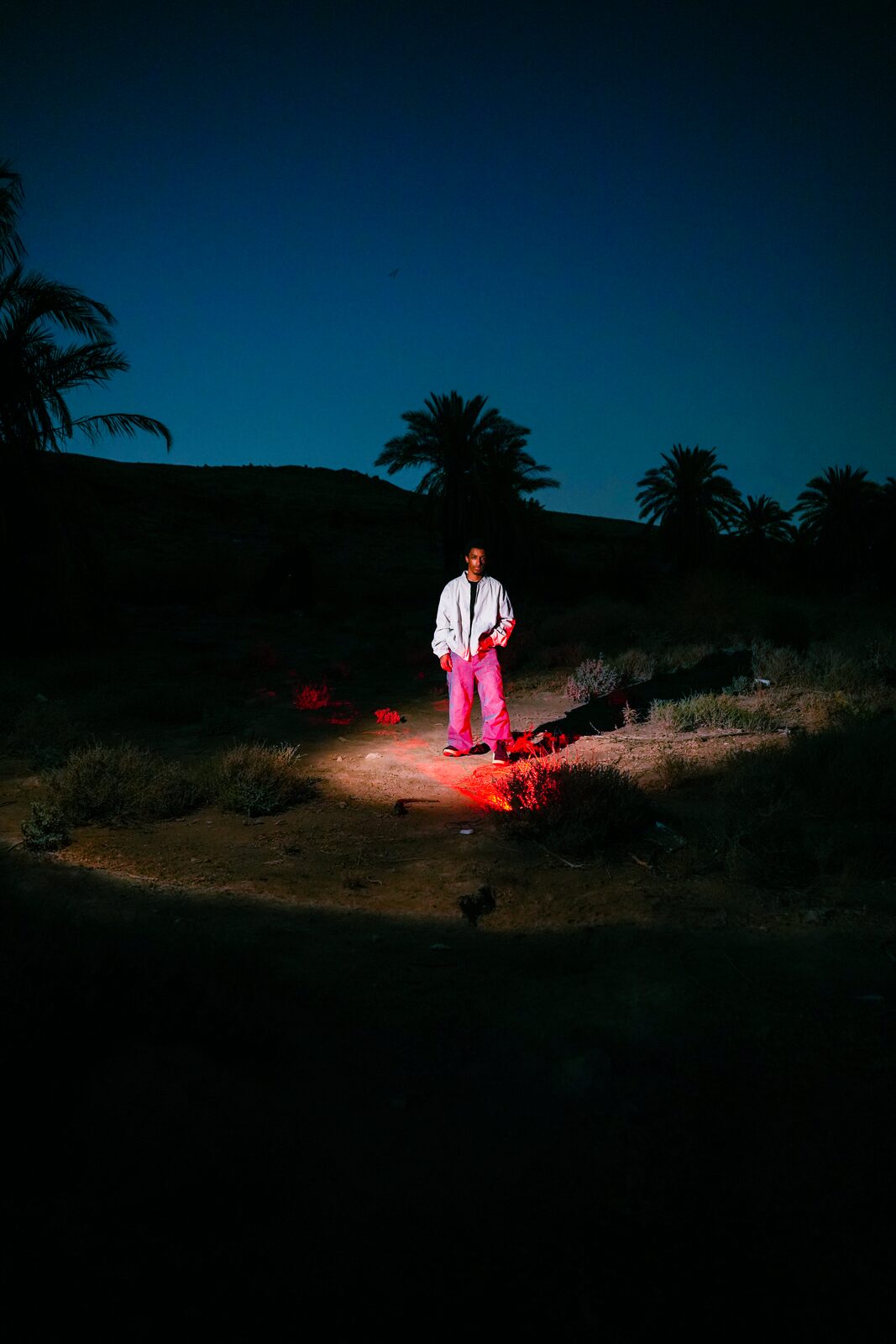 Figure in white and pink lit by red light in a dark palm grove — Salem Hamed Al Shammari (Traceofsalem) for ZELZAL