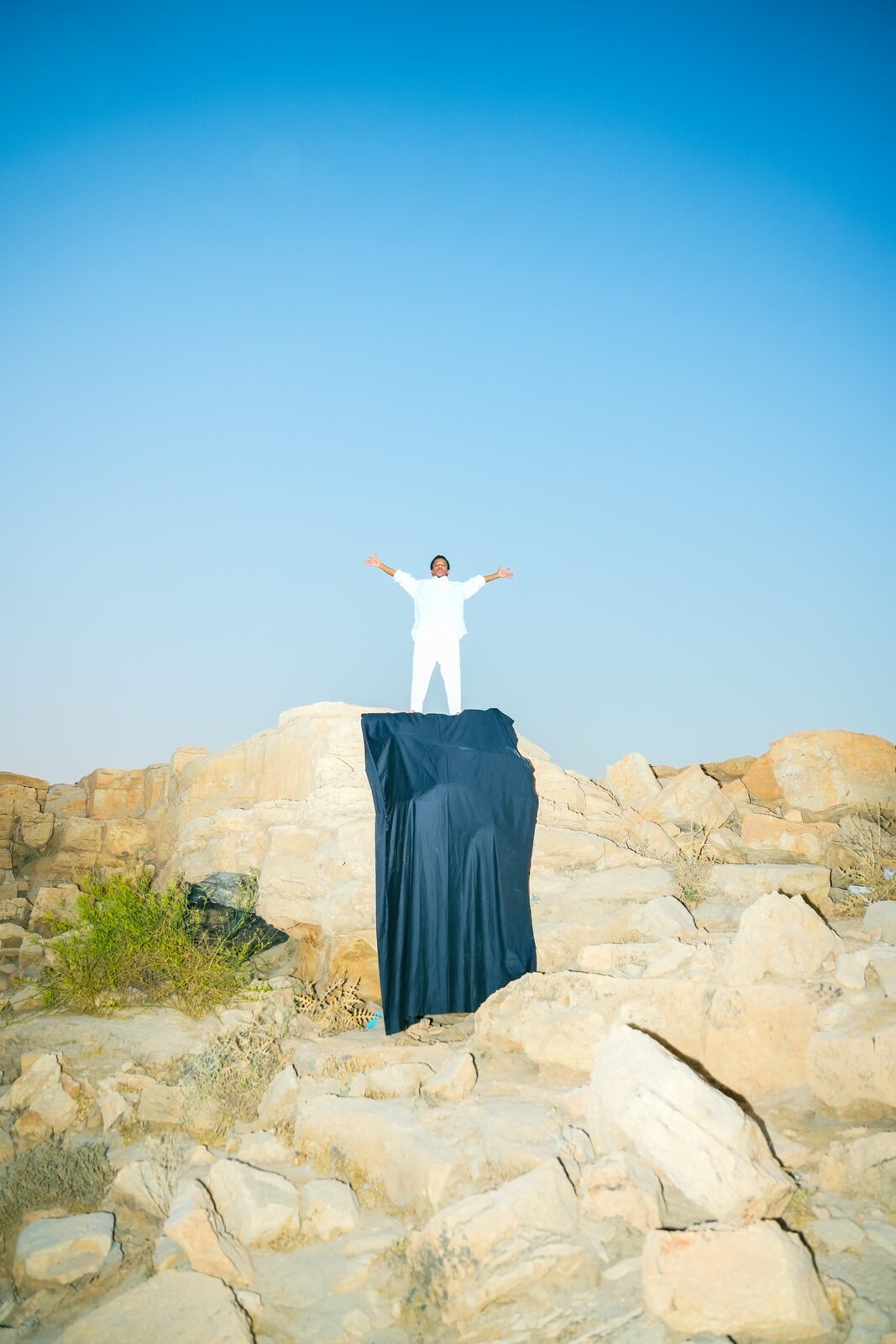 Figure in white with arms raised atop rocks, dark fabric draped below — Salem Hamed Al Shammari (Traceofsalem) for ZELZAL