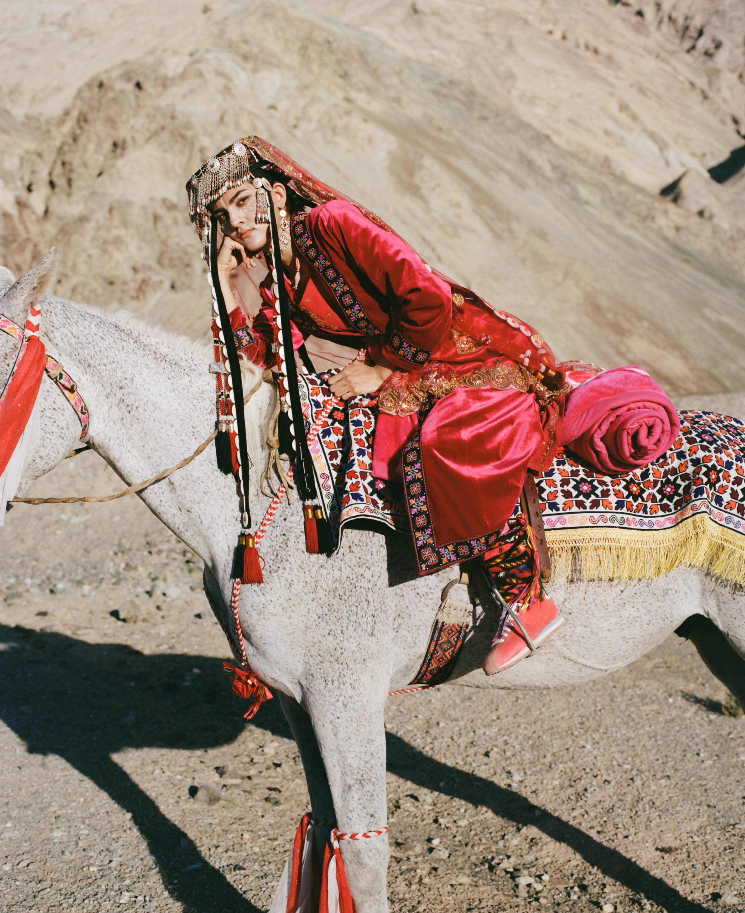 A woman in red velvet and a silver headdress rides side-saddle on a grey horse in desert mountains